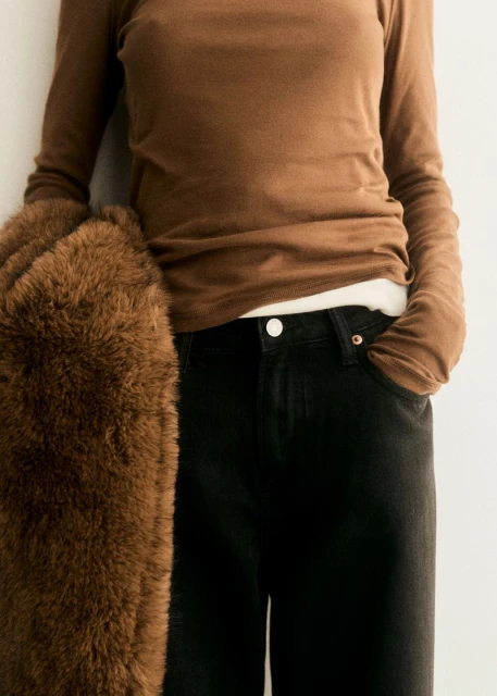 A person in a brown long-sleeve top and dark jeans, holding a brown faux fur jacket. Casual fashion shot with neutral background.