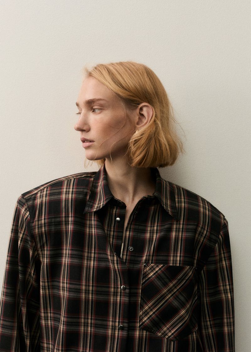 Model Tess Breeden wearing an oversized dark plaid button-up shirt against a neutral off-white background.