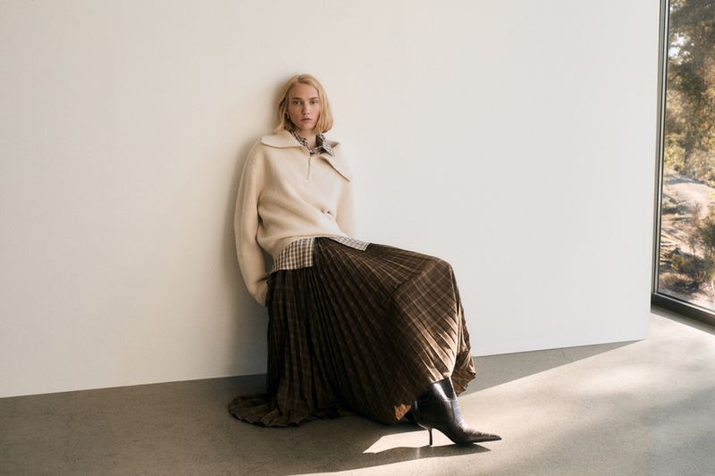 A fair-skinned woman in a beige sweater and brown plaid pleated skirt leans against a white wall indoors near a window.