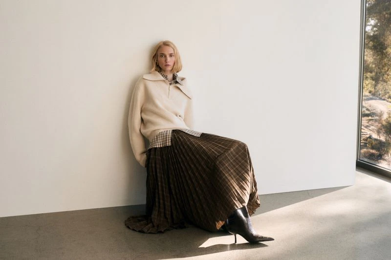 A fair-skinned woman in a beige sweater and brown plaid pleated skirt leans against a white wall indoors near a window.