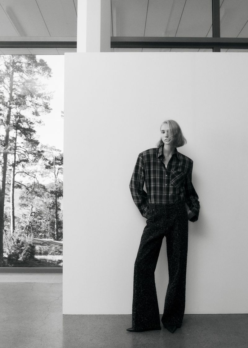 Black and white portrait of a slender fashion model in a plaid shirt and wide leg pants striking a pose against a white wall.