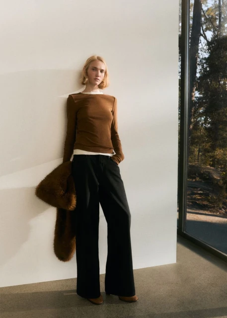 A woman wearing a brown long-sleeve shirt and black pants poses next to a window, with natural lighting and a fur shawl.