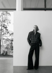Model in dark plaid shirt and black trousers leaning against a white partition wall with trees visible through a window.