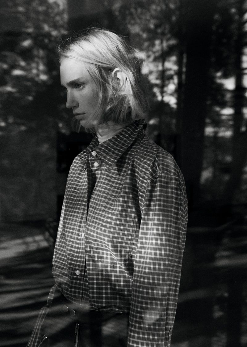 Model wearing a plaid shirt against a dark, out-of-focus background with natural light filtering through trees