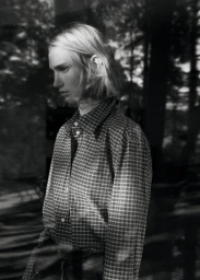 Model wearing a plaid shirt against a dark, out-of-focus background with natural light filtering through trees