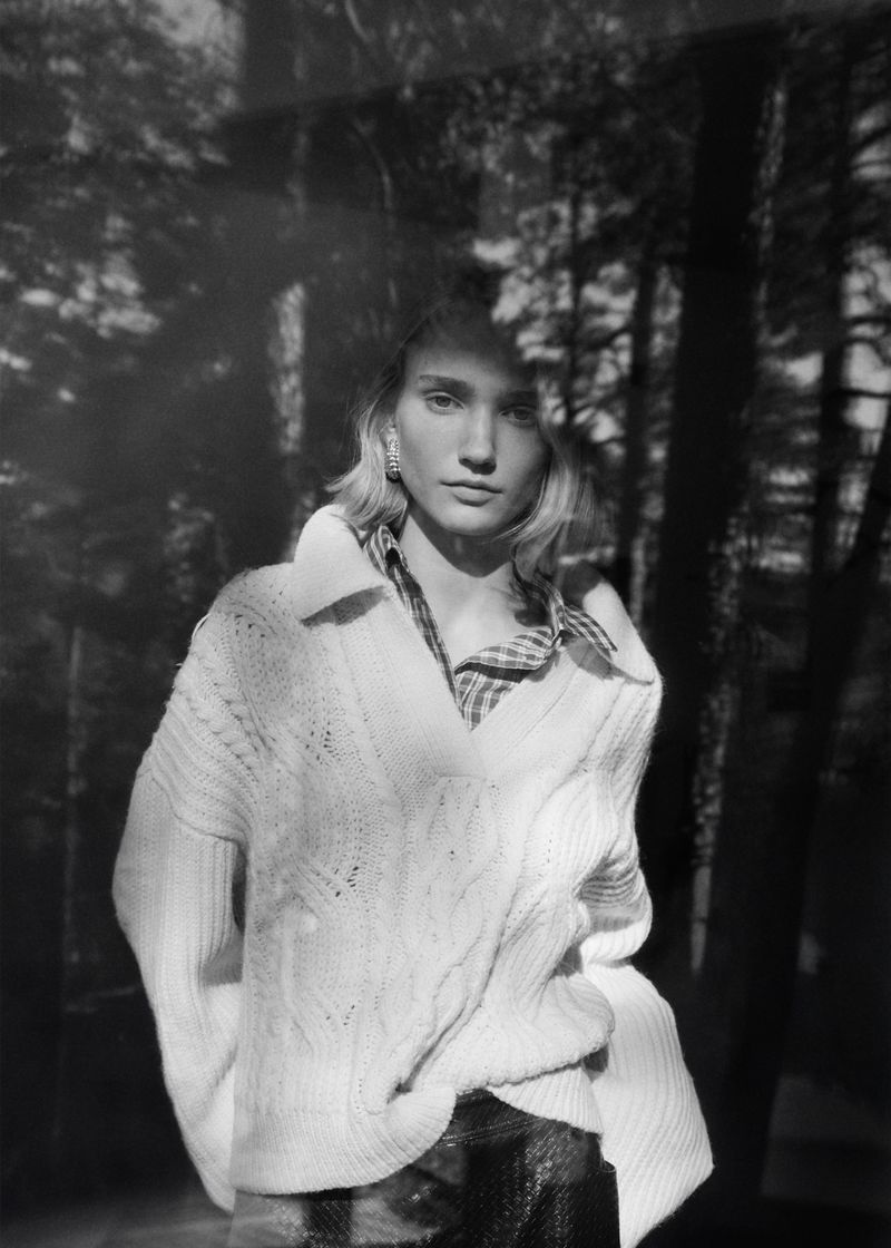 Model in a white knit sweater standing outdoors against a background of dark trees with layered double exposure tree reflections.