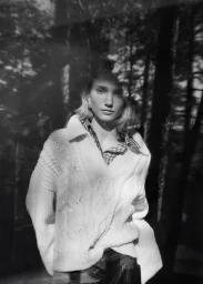 Model in a white knit sweater standing outdoors against a background of dark trees with layered double exposure tree reflections.