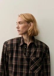 Model Tess Breeden wearing an oversized dark plaid button-up shirt against a neutral off-white background.