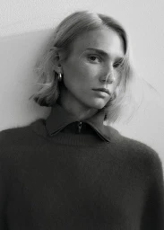 Portrait of a blonde woman wearing a textured knit sweater with a collared zip top against a plain light wall.