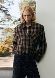 Model in plaid shirt standing indoors near a large window showcasing exterior trees and water
