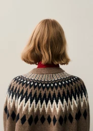 Back view of a model with a wavy blonde bob wearing a brown geometric knit Fair Isle sweater with a red collar on an off-white background.