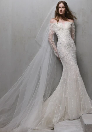 A woman in an off-the-shoulder, long-sleeved, beaded wedding dress and a long veil poses against a mottled grey wall in a studio.