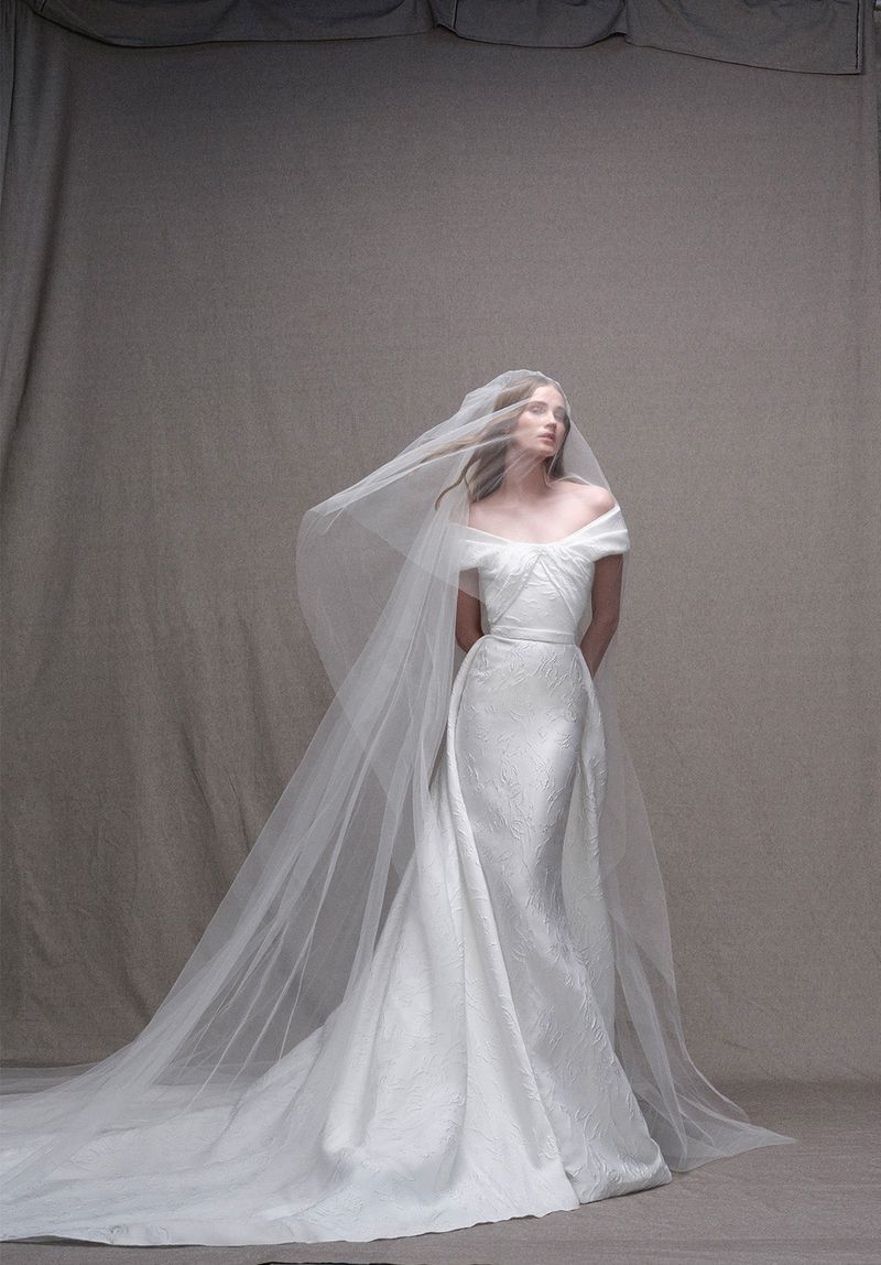Woman in an off-the-shoulder white textured gown with a long white veil poses in a studio, showcasing bridal fashion and style.