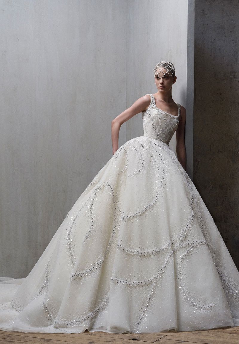 A woman poses in a large, formal white gown with glittering detail, styled with an ornate headpiece in a minimalist indoor setting.