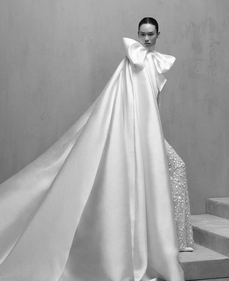Monochrome portrait of a fashion model in a satin gown with oversized bow detail and a flowing train, against a plain backdrop.