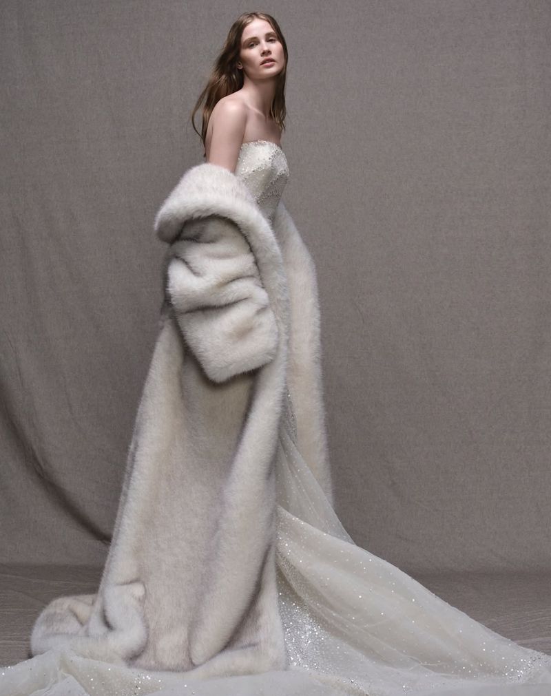 Elegant woman in an off-the-shoulder wedding dress with fur coat, standing against a neutral backdrop in a studio setting.