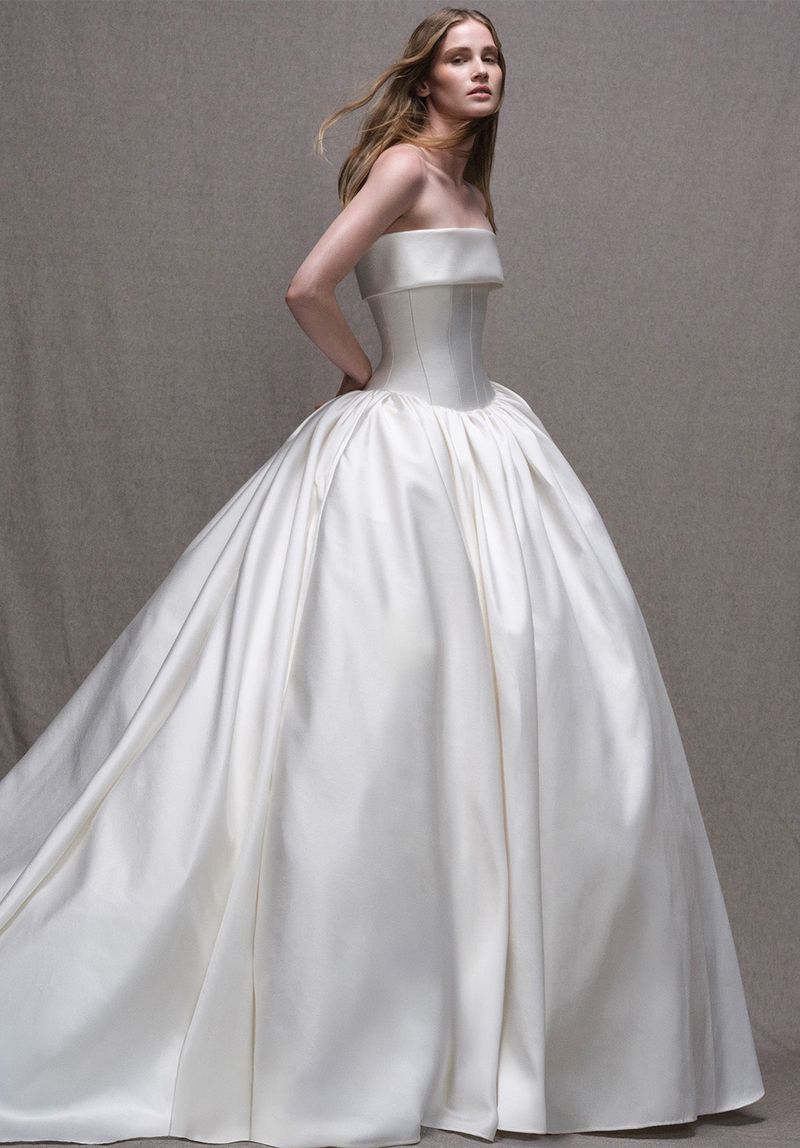 A fair-skinned woman models a strapless, white satin ballgown in a studio with a neutral background, looking directly at the camera.