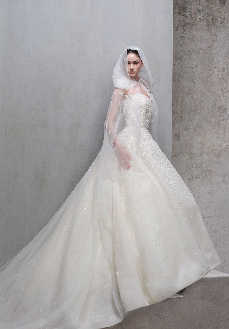 A fair-skinned woman in an elegant white couture bridal gown and veil poses against a minimalist grey backdrop in a fashion editorial.
