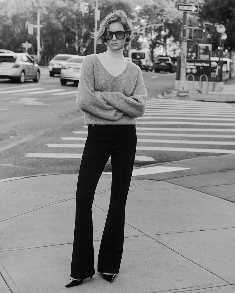 Black and white portrait of an adult female model in stylish sunglasses, v-neck sweater, and black bell-bottom pants on city street.