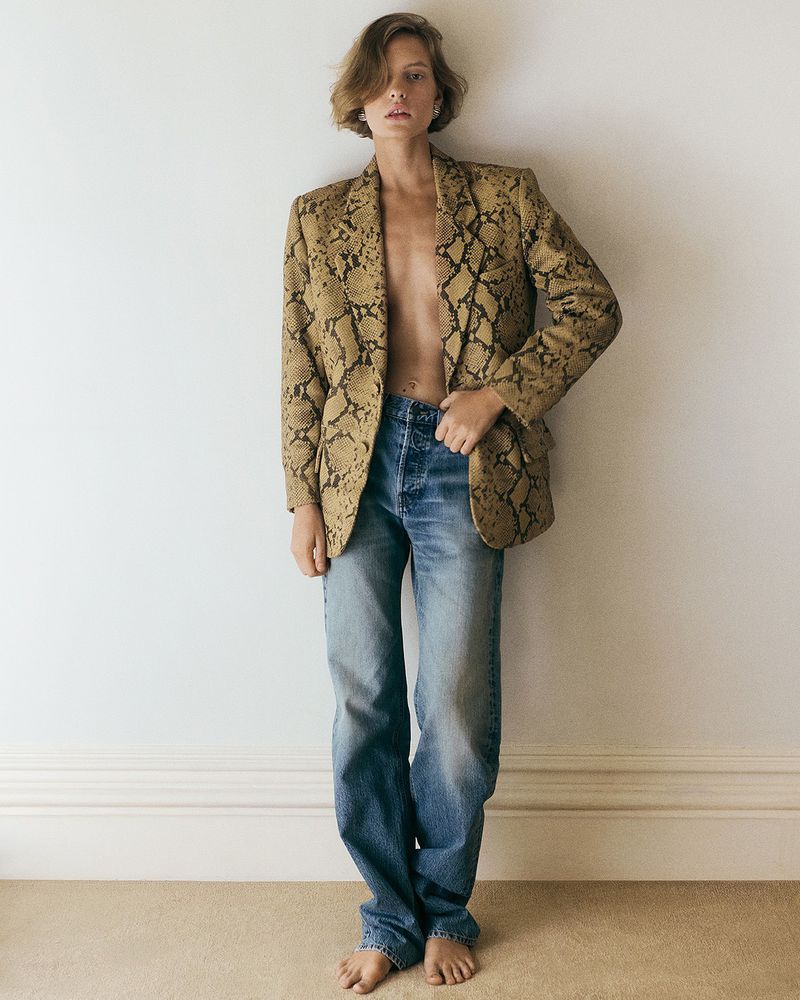 Barefoot woman poses in blue jeans and a snake-print blazer against a white wall, bathed in soft natural light for a fashion editorial.