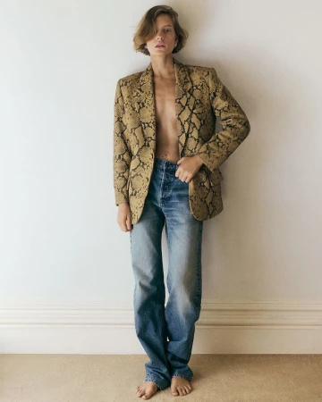 Barefoot woman poses in blue jeans and a snake-print blazer against a white wall, bathed in soft natural light for a fashion editorial.