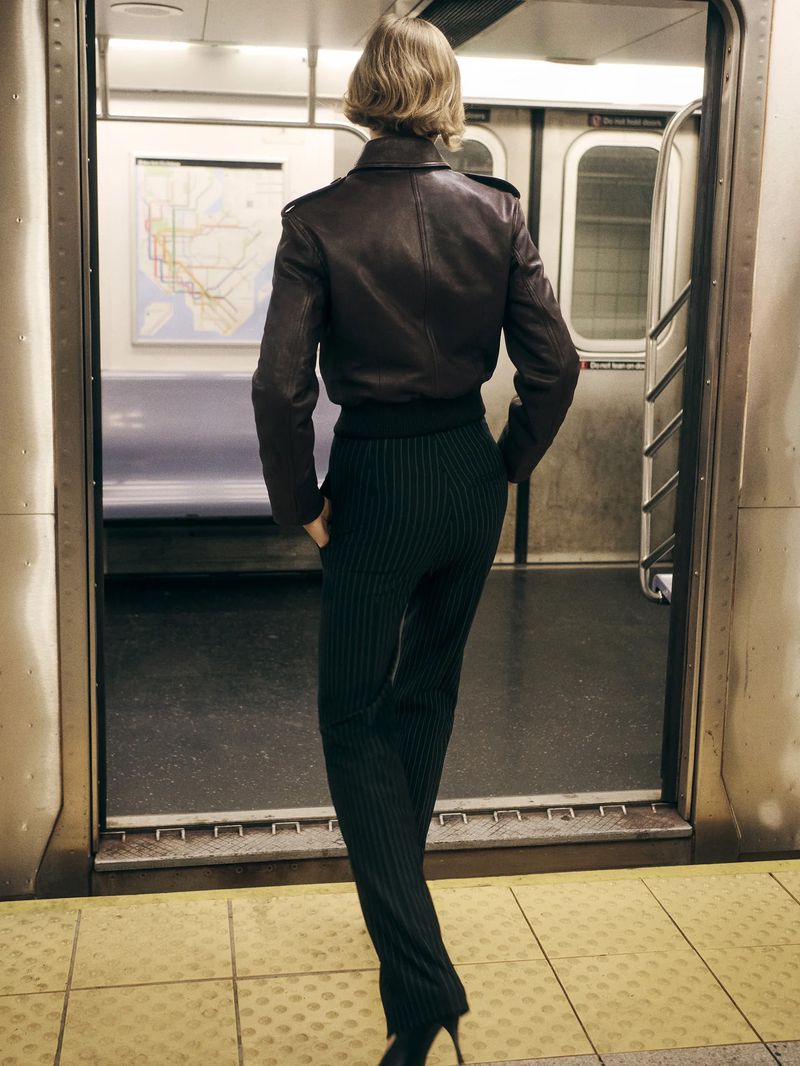 Back view of an adult female in a brown jacket and pinstripe pants standing in a subway car with a map in the background.