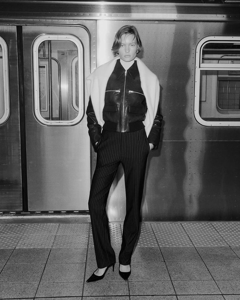 Black and white portrait of a woman in a leather jacket and striped pants posing in front of a subway train; shot at eye level.