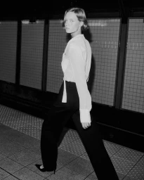 Model walking on a tiled subway platform wearing a white blouse and dark trousers for FRAME The Trouser Program.