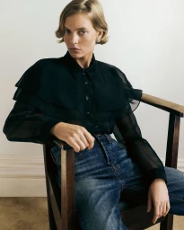 Model wearing a sheer black blouse and blue jeans seated in a wooden chair against a plain light wall.