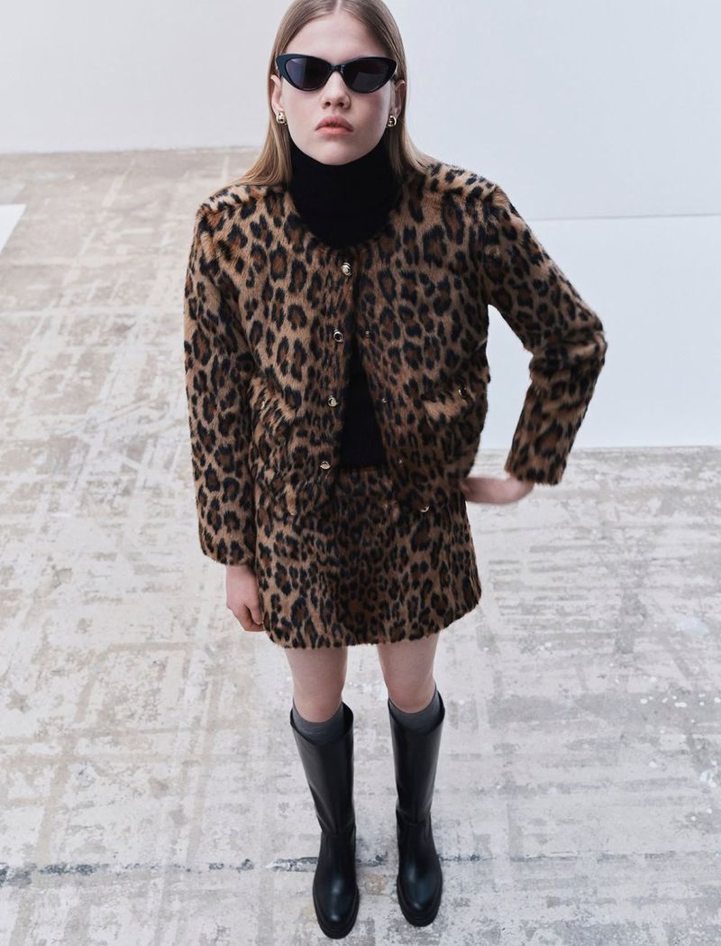 A woman in a leopard print jacket and skirt, black turtleneck, sunglasses, and boots stands in a bright studio. Fashion shot.