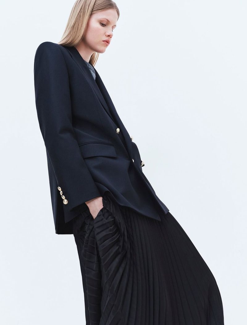 Model Sofi Damzina wearing an oversized black blazer with gold buttons and a pleated black skirt against a white studio background.