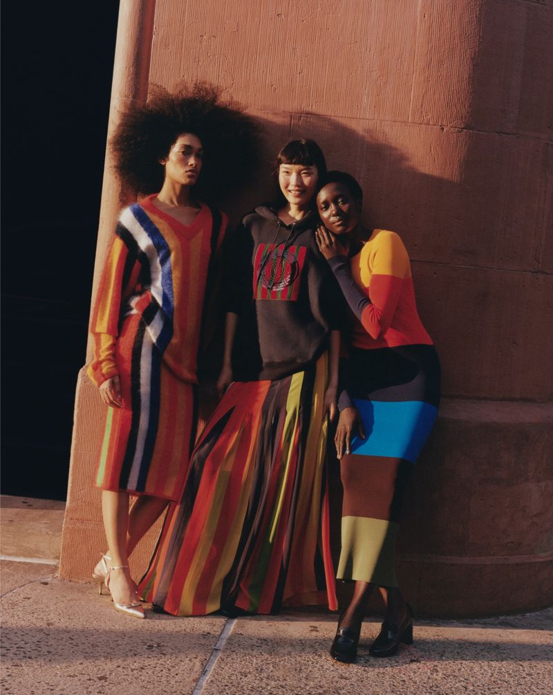 Three women model color-blocked knitwear dresses in an outdoor setting with a neutral wall and dark background, showcasing fashion and style.