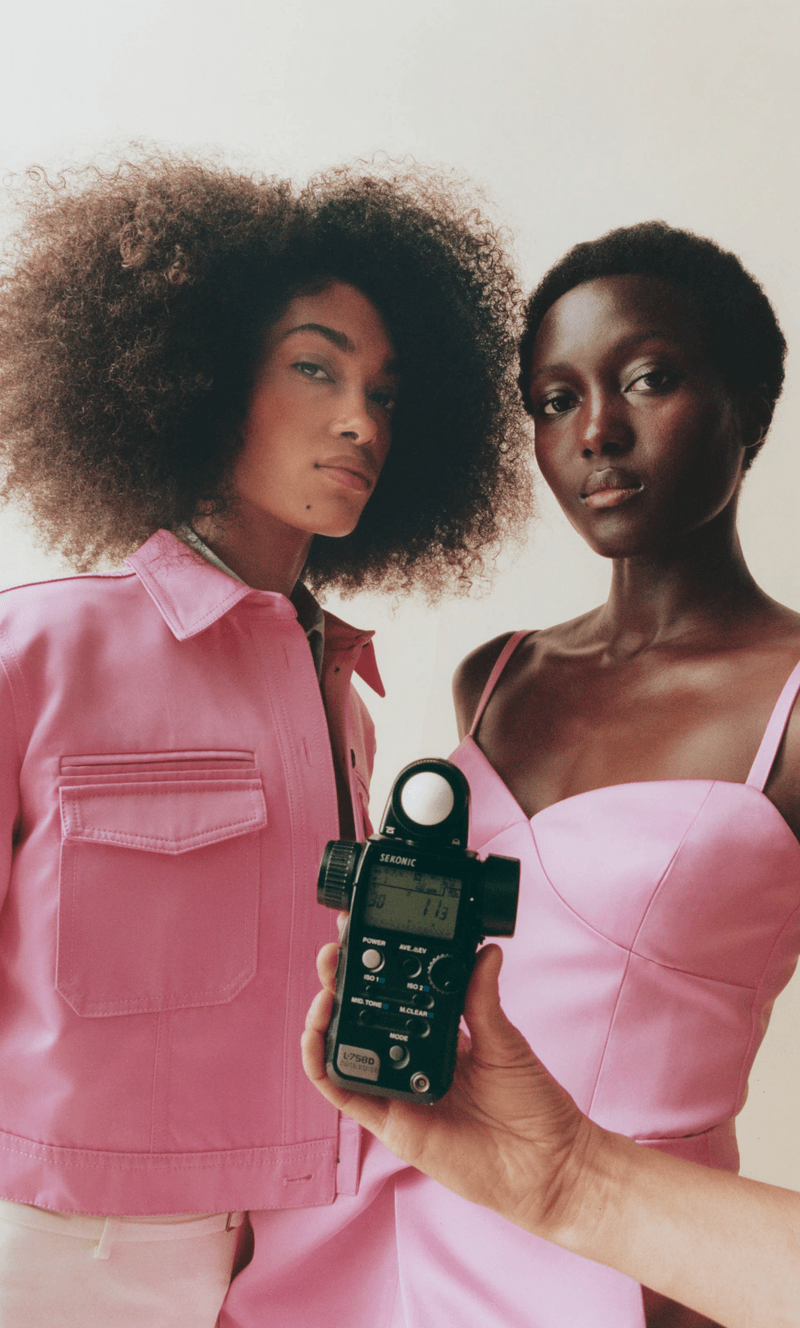 Two Black female models pose in pink clothing with a light meter held in the foreground against a neutral backdrop.