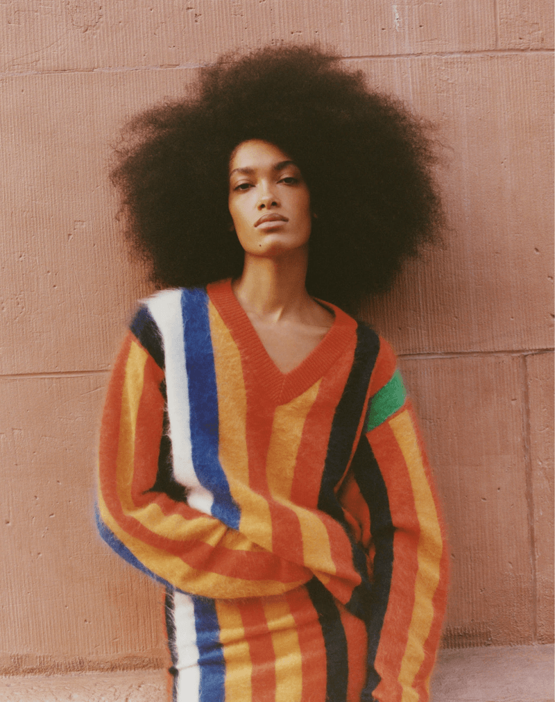 Adult woman with afro poses in front of a wall wearing a multicolor striped sweater. Portrait shot, eye-level angle, studio lighting.