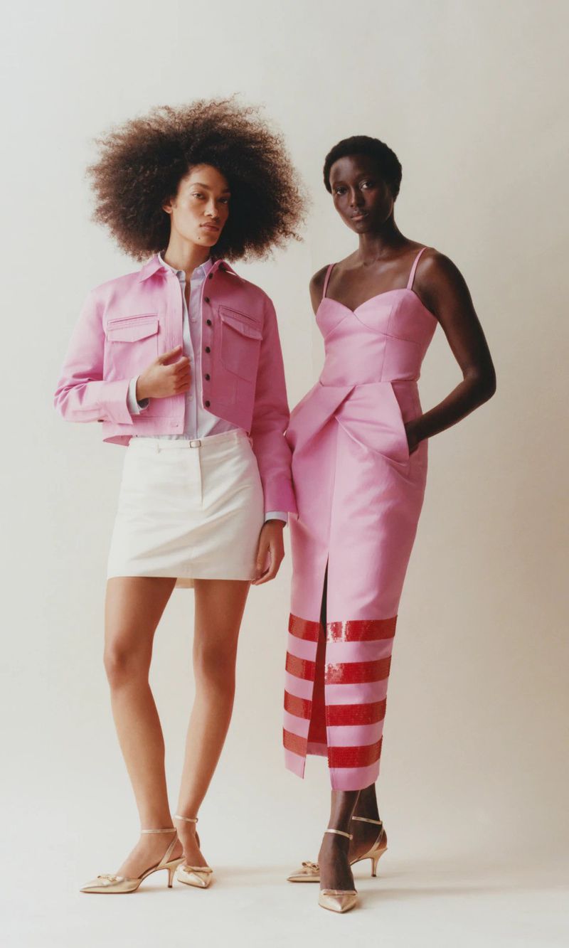 Two female fashion models pose in a studio, one wearing a pink jacket and skirt, the other a pink dress with red accents.