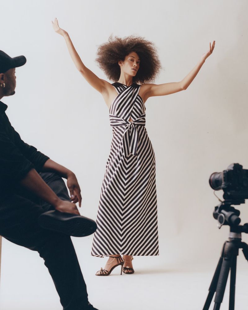 Fashion photo shoot: a Black adult female model in a striped dress poses with arms raised, while a photographer sits in the foreground.