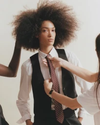 Model with voluminous afro hairstyle wearing a white shirt and black vest being adjusted by stylists in a studio setting