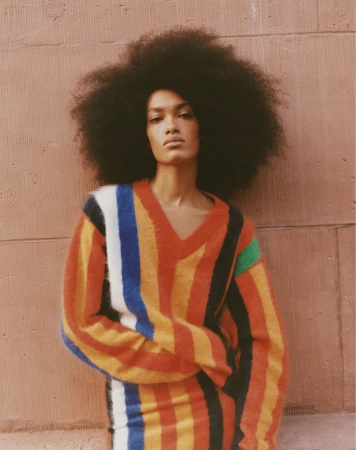 Model with large afro hair wearing a colourful striped V-neck sweater leaning against a textured brown brick wall.