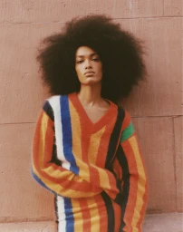 Model with large afro hair wearing a colourful striped V-neck sweater leaning against a textured brown brick wall.