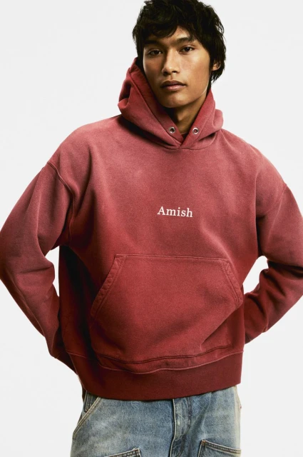 Man in a red-toned hooded sweatshirt with 'Amish' printed on the chest and blue jeans, posing in front of a plain background.