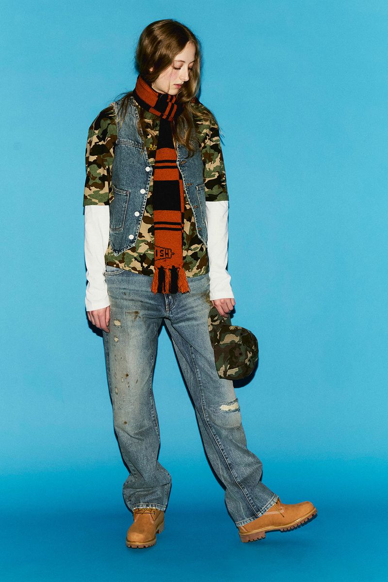Fashionable woman in camouflage shirt, denim vest, scarf, and light blue jeans, holding cap in a studio setting against blue backdrop.