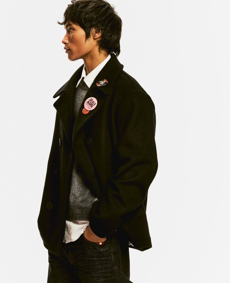 Profile shot of a young adult man wearing a black pea coat over a gray sweater, white collared shirt, and dark denim jeans.