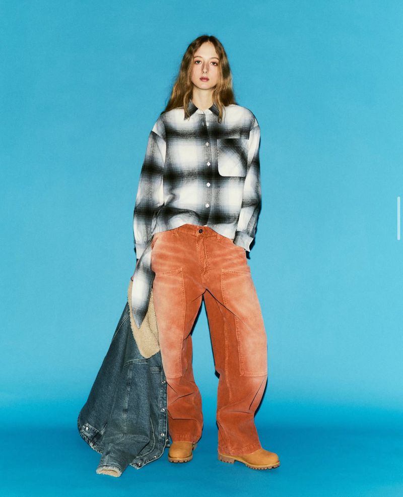 A young woman poses in a studio wearing a plaid shirt, corduroy pants, and tan work boots against a bright blue background.