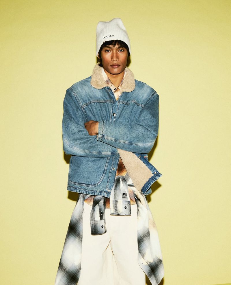 Man in denim jacket and beanie posing against a yellow wall. Fashionable outerwear and casual style in a studio setting.