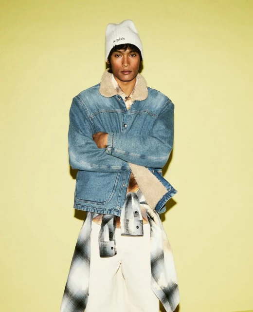 Man in denim jacket and beanie posing against a yellow wall.  Fashionable outerwear and casual style in a studio setting.