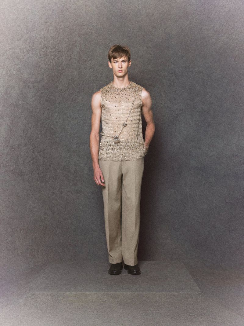 Model in a beige sleeveless embellished top and tailored beige trousers standing on a platform against a grey textured backdrop.