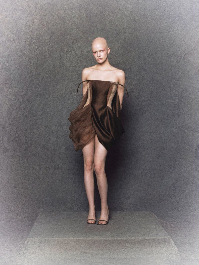 A bald woman with fair skin poses in a short, strapless brown dress with dark sandals, standing on a gray platform in a studio.