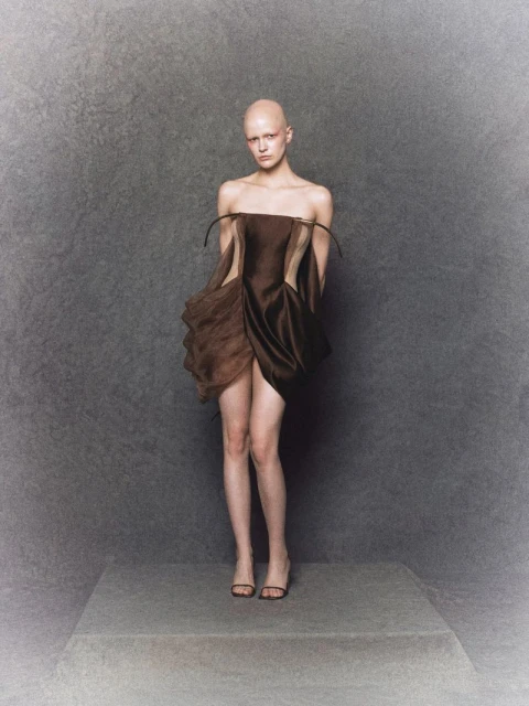 A bald woman with fair skin poses in a short, strapless brown dress with dark sandals, standing on a gray platform in a studio.