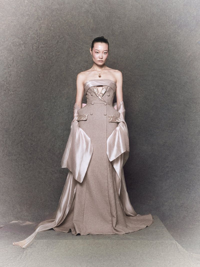 Woman in couture taupe gown with draped sleeves and train, standing in a neutral studio against a mottled grey background.