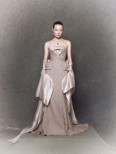 Woman in couture taupe gown with draped sleeves and train, standing in a neutral studio against a mottled grey background.